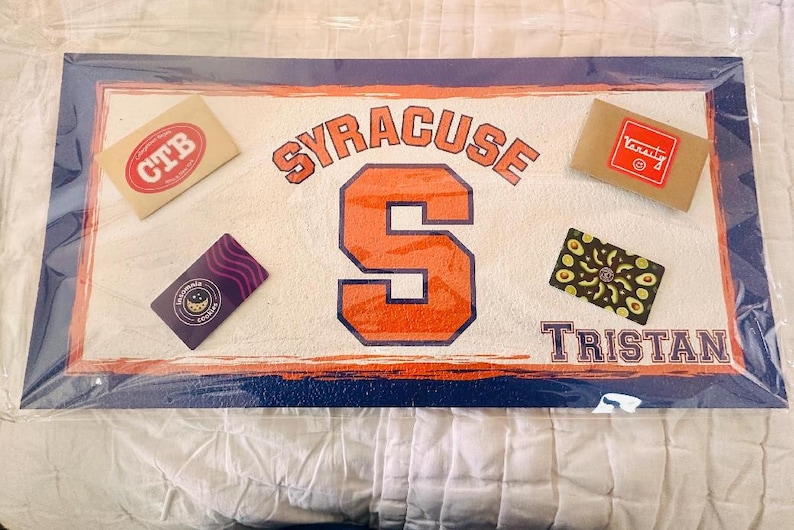 Op de afbeelding: Een rechthoekig decoratief item met een witte achtergrond, met het woord "SYRACUSE" in oranje en blauw, en een grote oranje "S". De naam "TRISTAN" staat onderaan. Kleine rechthoekige items met logo's zijn verspreid over het ontwerp.