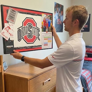 Op de afbeelding: Een zwart omrand prikbord met het Ohio State University-logo in rood en wit. Het bord bevat een familiefoto, een kalender en diverse kaarten. Het bord wordt voor een houten dressoir gehouden.