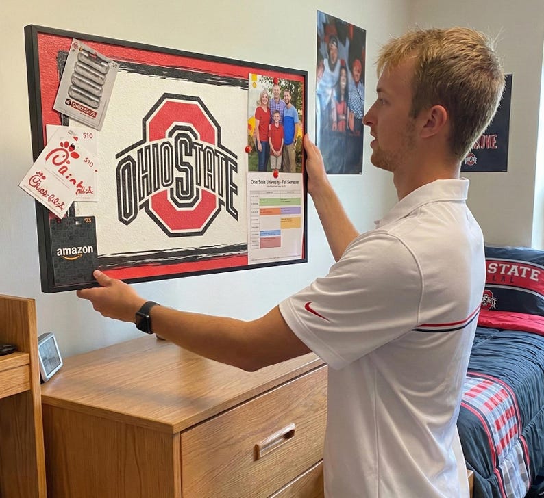 Op de afbeelding: Een zwart omrand prikbord met het Ohio State University-logo in rood en wit. Het bord bevat een familiefoto, een kalender en diverse kaarten. Het bord wordt voor een houten dressoir gehouden.
