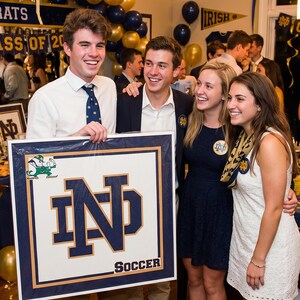 Op de afbeelding: Een groep mensen die glimlachen en poseren met een ingelijste poster. De poster toont het Notre Dame-logo en het woord "SOCCER". De achtergrond bevat gouden en blauwe ballonnen en banners.