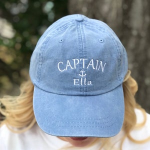 May include: A blue denim baseball cap with white embroidery that reads "CAPTAIN" with an anchor symbol and the name "Ella".
