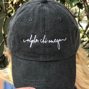 Puede incluir: Una gorra de béisbol negra con texto bordado en blanco que dice "alpha chi omega"