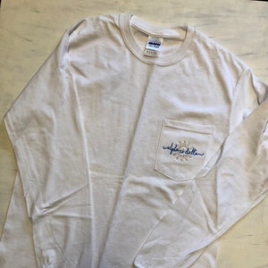 Puede incluir: Camiseta blanca de manga larga con un bolsillo en el pecho izquierdo. El bolsillo tiene un pequeño sol bordado y el texto "alpha delta" en azul.