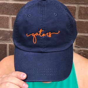K&ouml;nnte beinhalten: Eine marineblaue Baseballkappe mit einer orangefarbenen Stickerei, auf der "gators" steht.