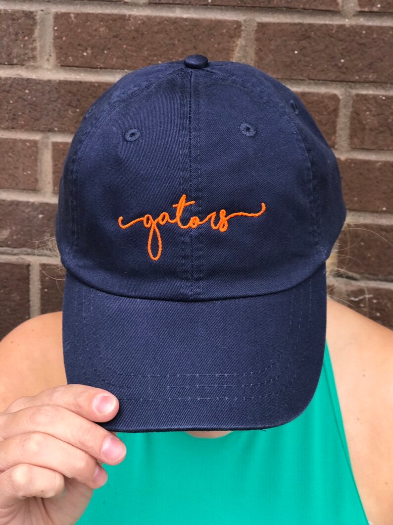 K&ouml;nnte beinhalten: Eine marineblaue Baseballkappe mit einer orangefarbenen Stickerei, auf der "gators" steht.