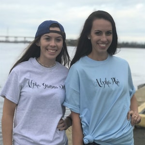 May include: Two women wearing t-shirts with sorority names. The woman on the left is wearing a grey t-shirt with "Alpha Gamma Delta" written in black script. The woman on the right is wearing a light blue t-shirt with "Alpha Phi" written in black script.