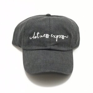 May include: A dark gray baseball cap with the embroidered text "hot mess express" in white script. The cap has a curved brim and a rounded crown. The hat is made of a soft, textured fabric.
