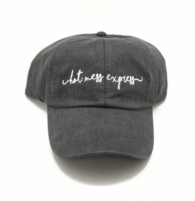 May include: A dark gray baseball cap with the embroidered text "hot mess express" in white script. The cap has a curved brim and a rounded crown. The hat is made of a soft, textured fabric.