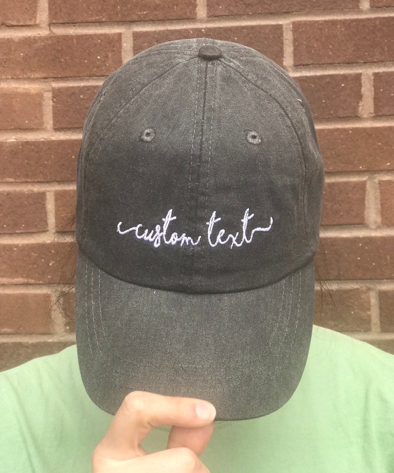 May include: A dark gray baseball cap with the embroidered text "custom text" in white script. The cap has a curved brim and a button on top. The hat is made of a soft, worn-looking fabric.