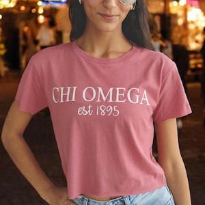 Puede incluir: Una camiseta rosa de manga corta y corte crop con texto blanco que dice "CHI OMEGA est 1895".