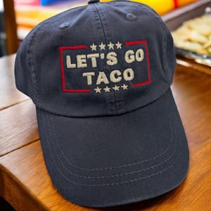 May include: Navy blue baseball cap with the embroidered text "LET'S GO TACO" in tan, framed by a red rectangle and stars. The cap is made of a durable fabric and has a curved brim.