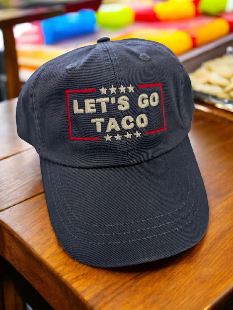 May include: Navy blue baseball cap with the embroidered text "LET'S GO TACO" in tan, framed by a red rectangle and stars. The cap is made of a durable fabric and has a curved brim.