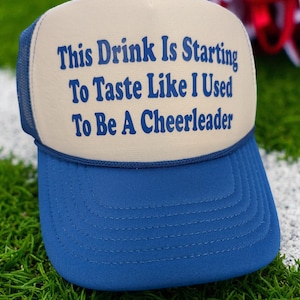 May include: A blue and tan trucker hat with the text "THIS DRINK Is Starting To Taste Like I Used To Be A Cheerleader" in blue lettering. The hat is on a green surface with white lines and red and white pom-poms in the background.