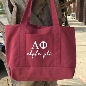 Puede incluir: Una bolsa de lona color burdeos con un bolsillo blanco que presenta las letras "AΦ" y las palabras "alpha phi" en una fuente de escritura.