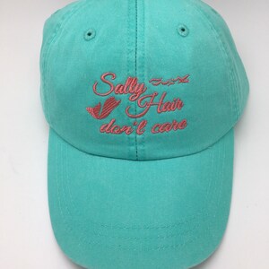 May include: A turquoise blue baseball cap with pink embroidered text that reads "Sally Hair don't care".