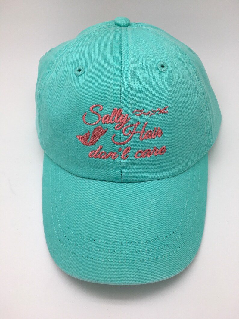 May include: A turquoise blue baseball cap with pink embroidered text that reads "Sally Hair don't care".
