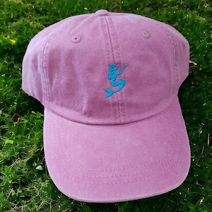 Puede incluir: Una gorra de b&eacute;isbol rosa con un dise&ntilde;o de sirena bordado en azul.
