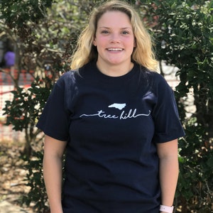May include: A woman wearing a navy blue t-shirt with a white outline of the state of North Carolina and the text "tree hill" underneath. She is standing in front of a green bush.