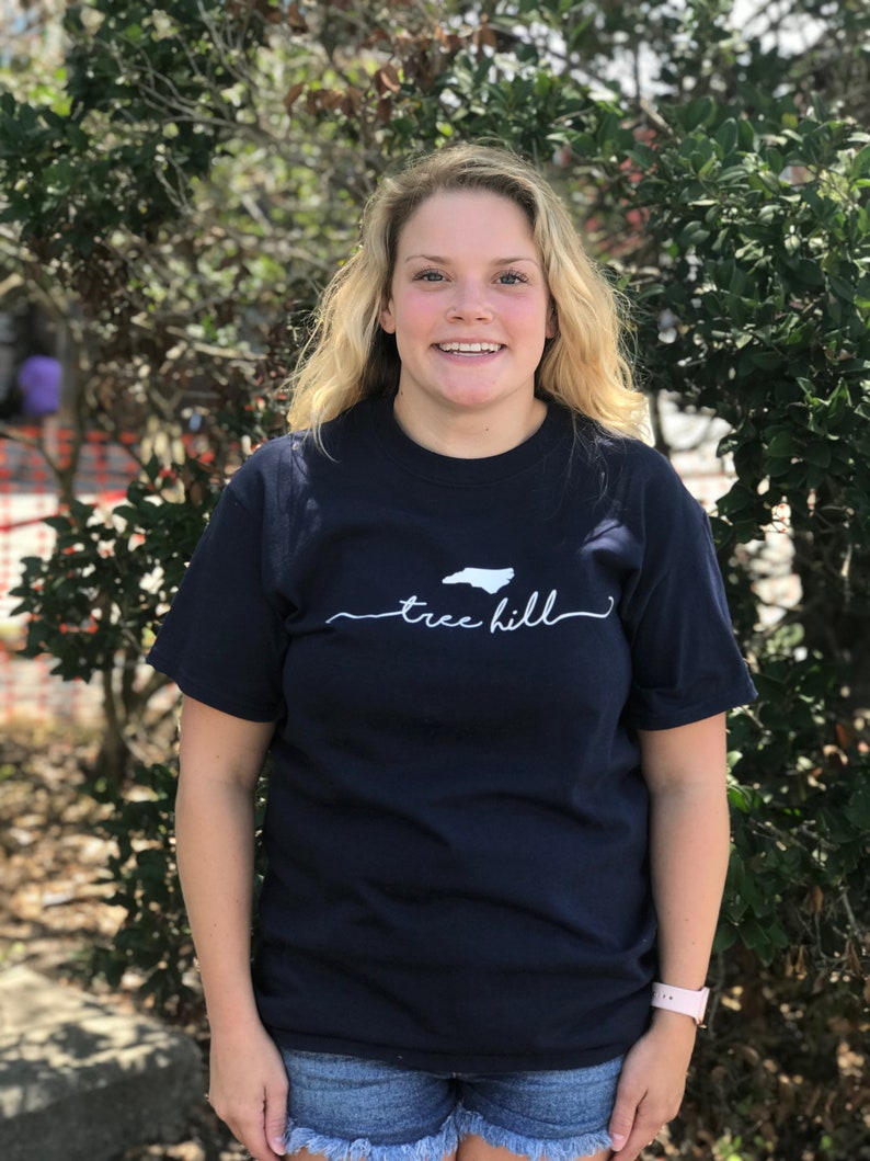 May include: A woman wearing a navy blue t-shirt with a white outline of the state of North Carolina and the text "tree hill" underneath. She is standing in front of a green bush.