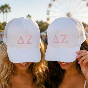 May include: Two white trucker-style baseball caps featuring a mesh back and a solid front panel. Each cap is embroidered with the Greek letters Delta and Zeta in a light pink thread centered on the front. The hats have a structured brim.