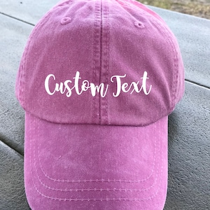 May include: A pink baseball cap with the text "Custom Text" embroidered in white on the front.