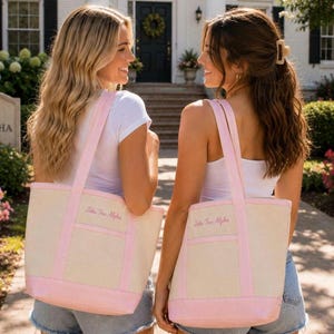 May include: A cream-coloured canvas tote bag with light pink trim, two shoulder straps, and a front pocket. The bag features the embroidered text Zeta Tau Alpha in pink script on the front panel, centred above the pocket.