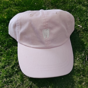 May include: A pink baseball cap with a white embroidered tooth on the front.
