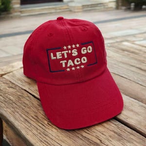 May include: A red baseball cap with the text "LET'S GO TACO" embroidered in white and outlined in blue. The cap has a curved brim and a rounded top. The hat is sitting on a wooden surface.