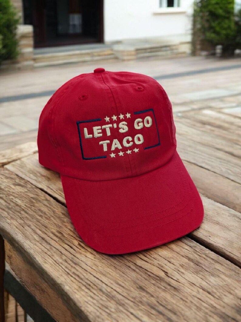 May include: A red baseball cap with the text "LET'S GO TACO" embroidered in white and outlined in blue. The cap has a curved brim and a rounded top. The hat is sitting on a wooden surface.