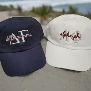 May include: Two baseball caps, one navy blue with white embroidery that says "delta gamma" and the other white with burgundy embroidery that says "alpha phi".