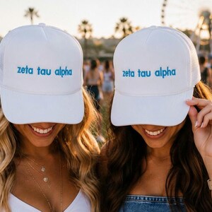 May include: Two white trucker hats featuring the text zeta tau alpha in blue lettering. Each hat has a structured front panel and a mesh back, with a curved brim. The design is identical on both hats, showcasing the blue text centered on the front.