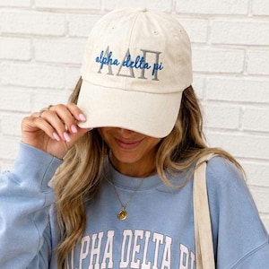 May include: A cream-coloured baseball cap featuring the embroidered Greek letters Alpha Delta Pi in silver thread, overlaid with the text alpha delta pi in blue cursive script. The cap has a structured crown, a curved brim, and ventilation eyelets.
