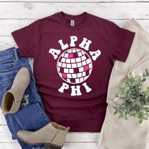 Op de afbeelding: Een maroon t-shirt met een witte afbeelding van een discobal en de tekst "ALPHA PHI" in een retro-lettertype.