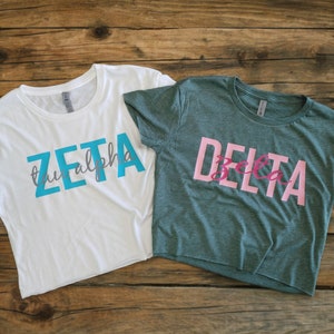 Op de afbeelding: Twee gecropte T-shirts, een wit met de tekst "ZETA tau alpha" in turquoiseblauw en grijs, en een groen met de tekst "DELTA Zeta" in roze.