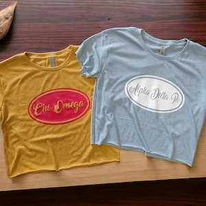 Puede incluir: Dos camisetas recortadas, una de color amarillo mostaza con un óvalo rojo y letras doradas que dicen "Chi Omega", y la otra azul claro con un óvalo blanco y letras grises que dicen "Alpha Delta Pi".
