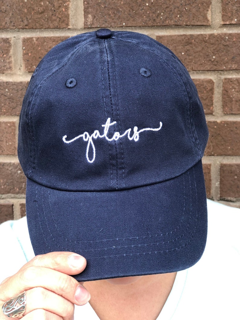 K&ouml;nnte beinhalten: Eine marineblaue Baseballkappe mit wei&szlig; gesticktem Text, der "gators" lautet.
