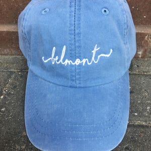 May include: A light blue baseball cap with the word "belmont" embroidered in white script across the front. The cap is made of a soft, textured fabric and features a curved brim and a button on top.