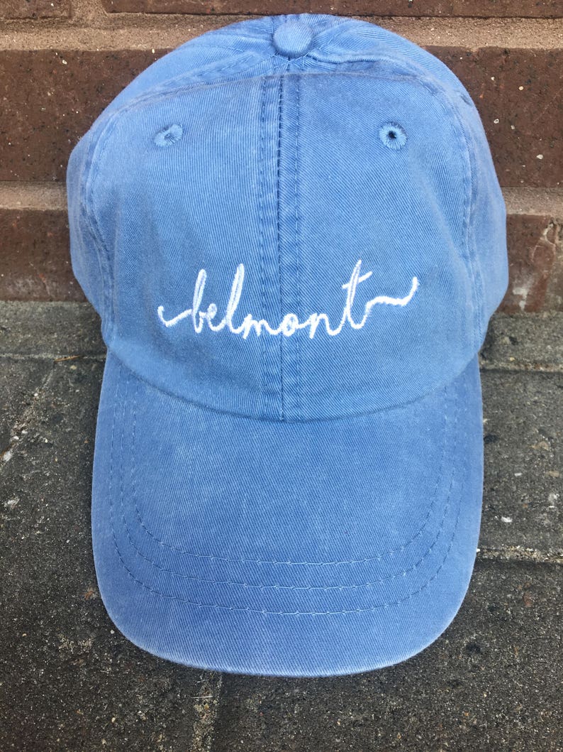May include: A light blue baseball cap with the word "belmont" embroidered in white script across the front. The cap is made of a soft, textured fabric and features a curved brim and a button on top.