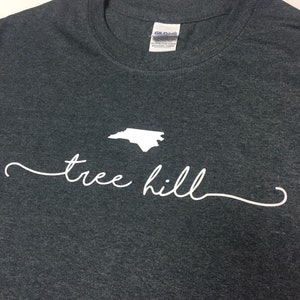 May include: A dark gray t-shirt with a white outline of the state of North Carolina and the text "tree hill" in white cursive.