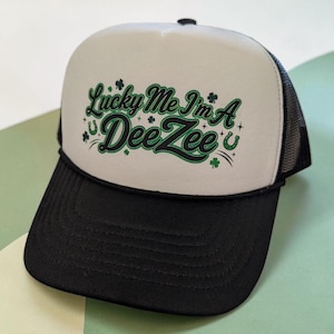 Op de afbeelding: Een wit met zwarte truckerpet met een zwarte klep en een mesh achterkant. Het voorpaneel toont groene en zwarte tekst met de tekst Lucky Me I'm A DeeZee, geaccentueerd met kleine groene klaver- en hoefijzerafbeeldingen en kleine sterretjes.