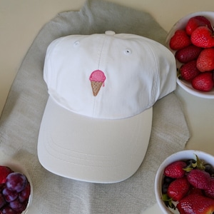 May include: White baseball cap with an embroidered pink ice cream cone on the front. The cap is on a light brown fabric surface with bowls of fruit nearby.