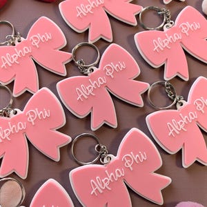 May include: Pink bow-shaped keyrings with the text "Alpha Phi" in white. The keyrings have a white border and silver key rings. The image shows multiple keyrings, suggesting a sorority or organization theme.