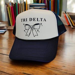 Puede incluir: Gorra de camionero blanca y azul marino con un lazo negro y el texto "TRI DELTA" en la parte delantera.