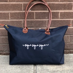 Puede incluir: Una bolsa de lona azul marino con asas de cuero marrón y cierre de cremallera. La bolsa tiene el texto "sigma sigma sigma" bordado en blanco en la parte delantera.