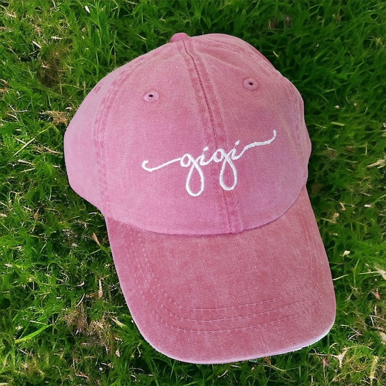 May include: A pink baseball cap with a white embroidered cursive word "gigi" on the front.