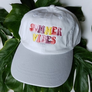 May include: White baseball cap with a colourful embroidered design that says "SUMMER VIBES" in a retro font.