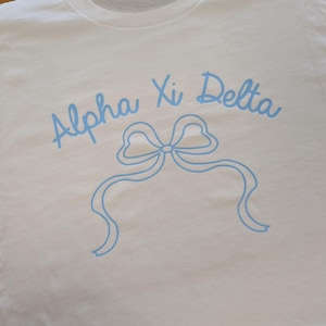 Puede incluir: Camiseta blanca con el texto azul claro "Alpha Xi Delta" sobre un gráfico de lazo azul claro. La camiseta está hecha de un material suave y tiene cuello redondo.