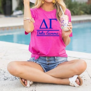 Op de afbeelding: Een roze T-shirt met een blauw Grieks letterontwerp met de tekst "ΔΓ Delta Gamma".