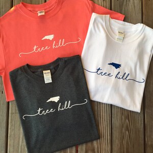 May include: Three t-shirts with the text "tree hill" and an outline of the state of North Carolina. The shirts are in coral, white, and dark grey.