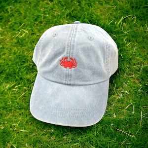 May include: A light blue denim baseball cap with a red embroidered crab on the front.
